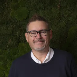 A person wearing a dark navy sweater over a white collared shirt and light brown pants stands against a textured green wall with moss-like plants. Hands are in pockets, and a watch is visible on the left wrist.