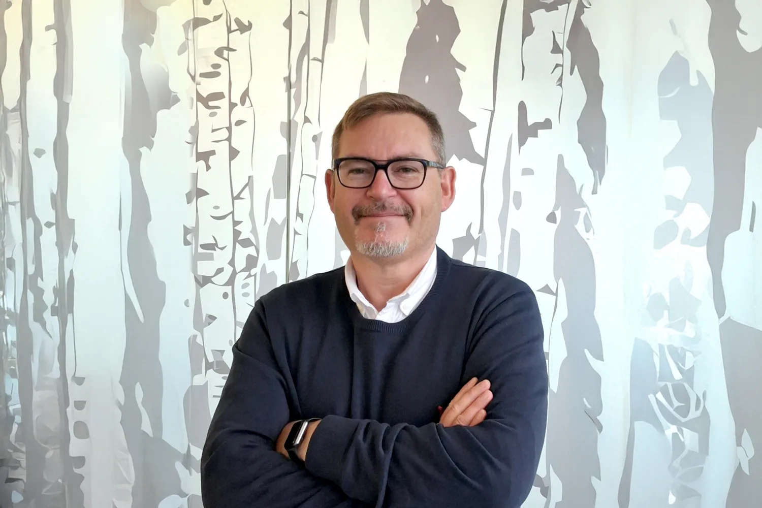 A person with short hair and glasses stands with arms crossed, wearing a dark sweater over a white collared shirt, in front of a mural depicting tall, slender trees in gray and white tones.