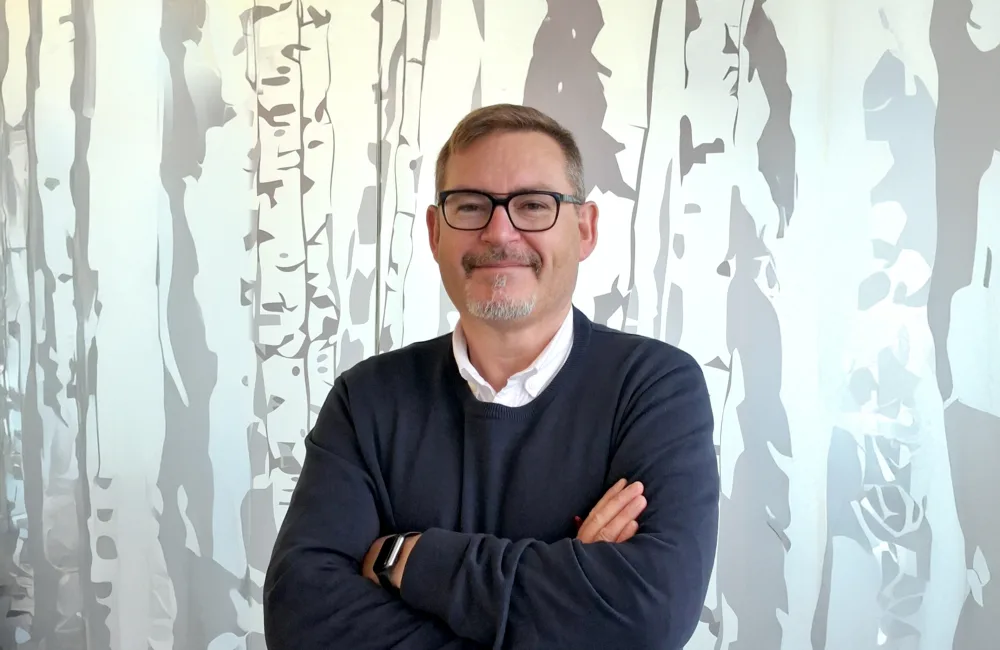 A person with short hair and glasses stands with arms crossed, wearing a dark sweater over a white collared shirt, in front of a mural depicting tall, slender trees in gray and white tones.