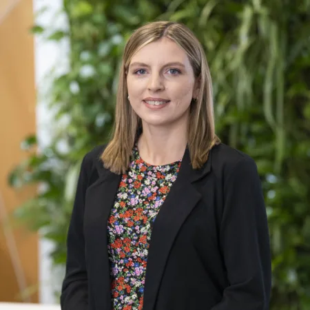 A person wearing a black blazer over a floral-patterned top stands indoors. The background features a green vertical plant wall and a section of light brown geometric panelling.
