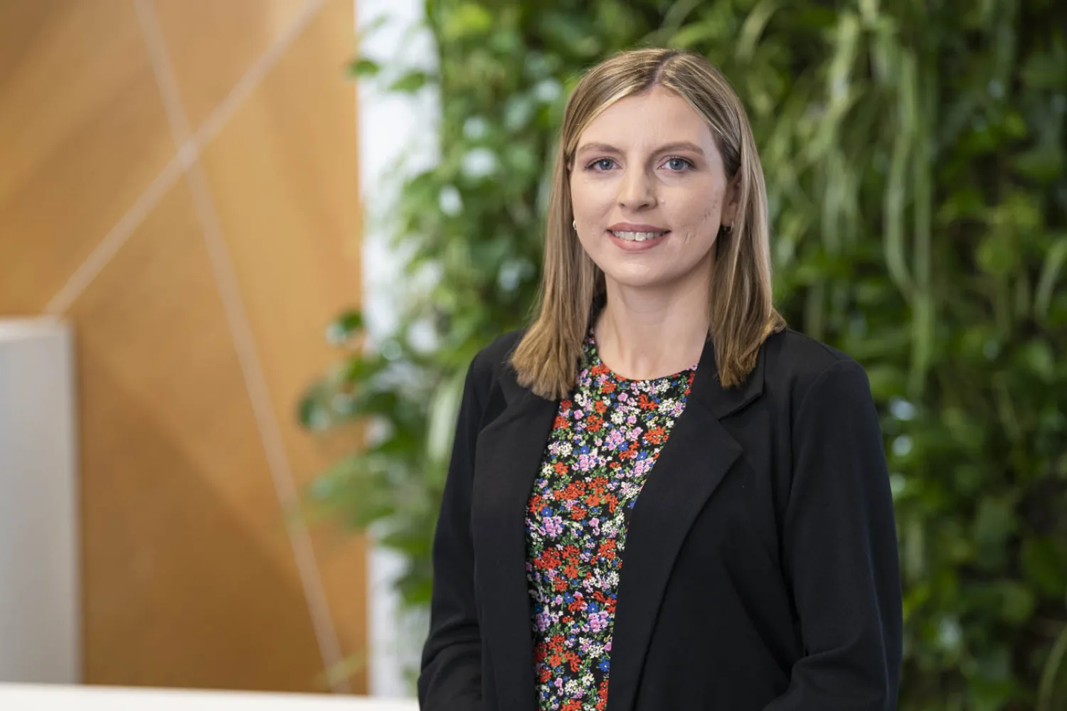 A person wearing a black blazer over a floral-patterned top stands indoors. The background features a green vertical plant wall and a section of light brown geometric panelling.