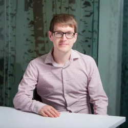 Stuart sat with his arm resting on a white desk in an open collar pale pink shirt and glasses in front of a frosted glass wall.