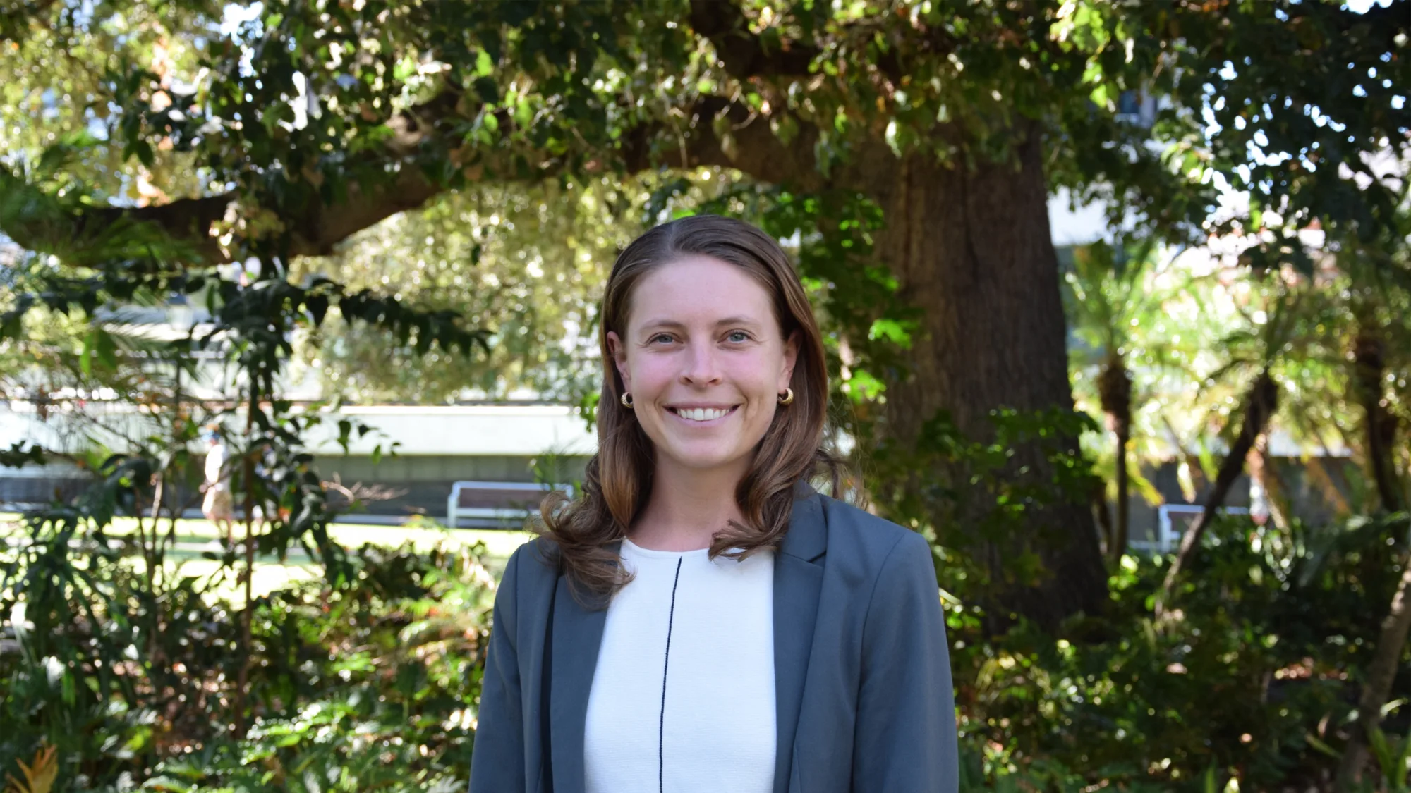 Anna Gannett wearing a light-colored top and a gray blazer standing outdoors in front of large tree and greenery, with benches and a building partially visible in the background.