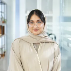 Ayesha Ahmed in light-colored garment with head covering standing indoors; background includes shelves with items, plants, and glass partitions in a modern, organized setting.