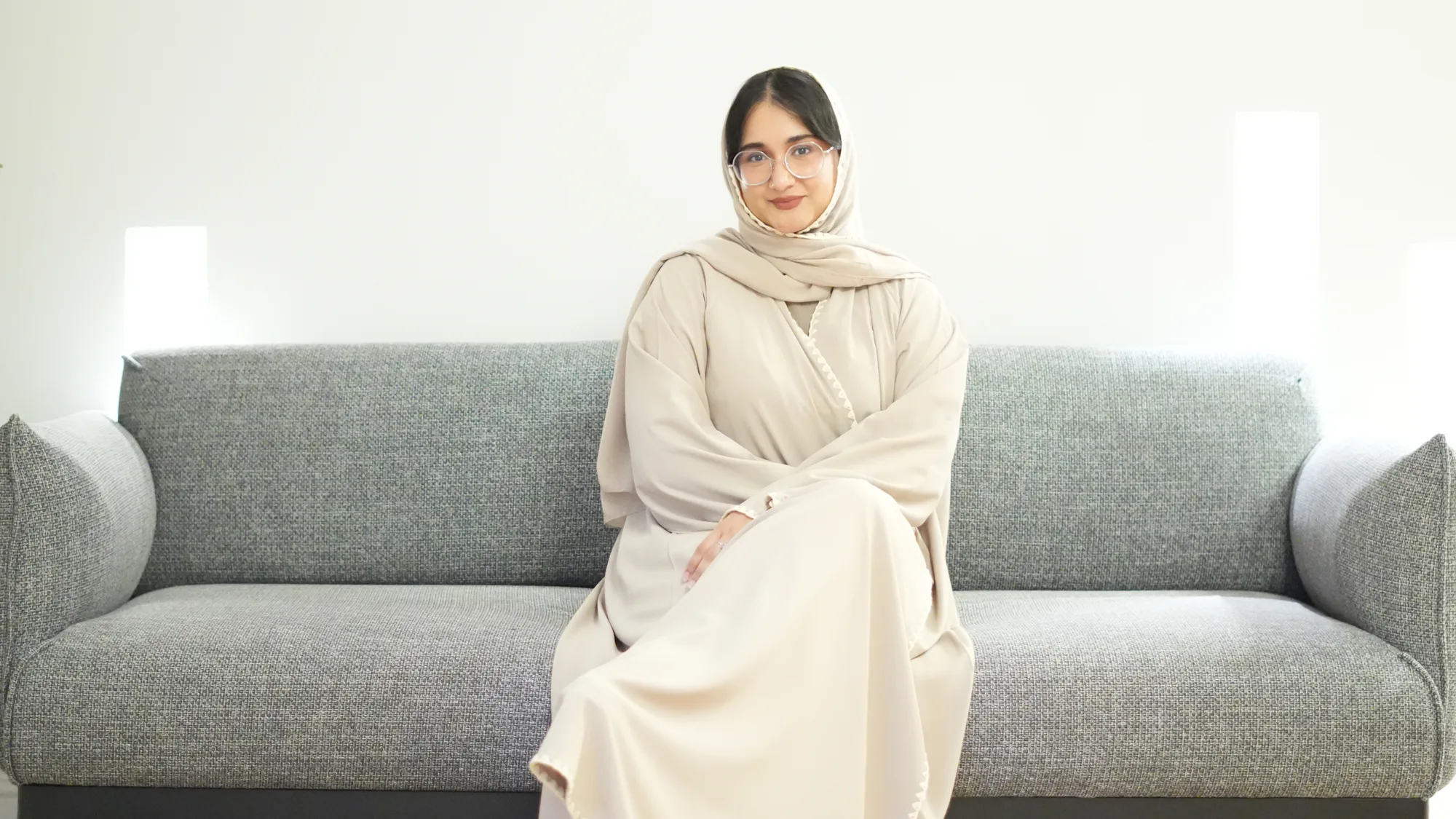 Ayesha Ahmed seated on a grey upholstered sofa wearing a light-colored flowing garment; hands resting on lap; background features two vertical light fixtures in a minimalist interior setting.