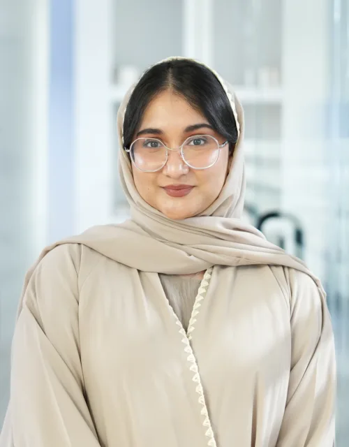 Ayesha Ahmed in light-colored garment with head covering standing indoors; background includes shelves with items, plants, and glass partitions in a modern, organized setting.