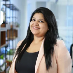 A person with long dark hair stands in an office setting, wearing a light-coloured blazer over a black top. Behind them are shelves with plants, glass walls, doors, and office furniture.