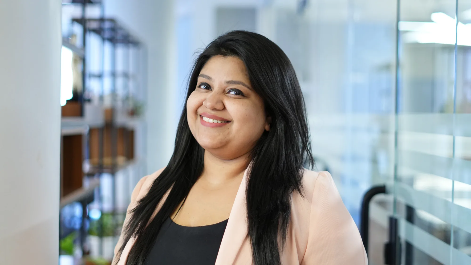 A person with long dark hair stands in an office setting, wearing a light-coloured blazer over a black top. Behind them are shelves with plants, glass walls, doors, and office furniture.