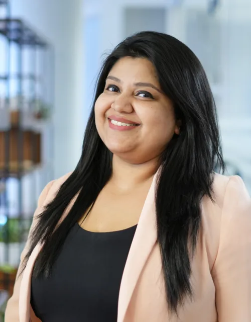 A person with long dark hair stands in an office setting, wearing a light-coloured blazer over a black top. Behind them are shelves with plants, glass walls, doors, and office furniture.