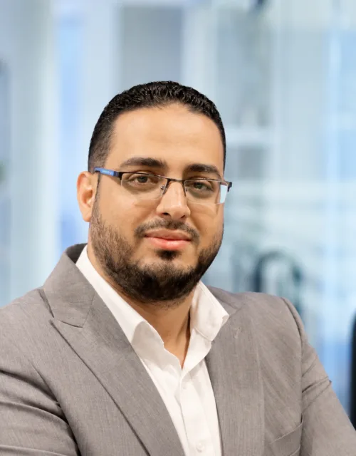Portrait of a person wearing rectangular glasses, a light grey blazer, and a white shirt, facing the camera. Background shows a softly blurred modern office interior with shelves and glass partitions.