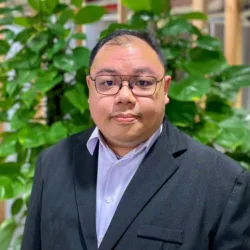 Man wearing a dark suit jacket and light dress shirt stands in front of a wooden slat wall with green leafy plants. Red pipes and industrial elements are visible in the indoor background.