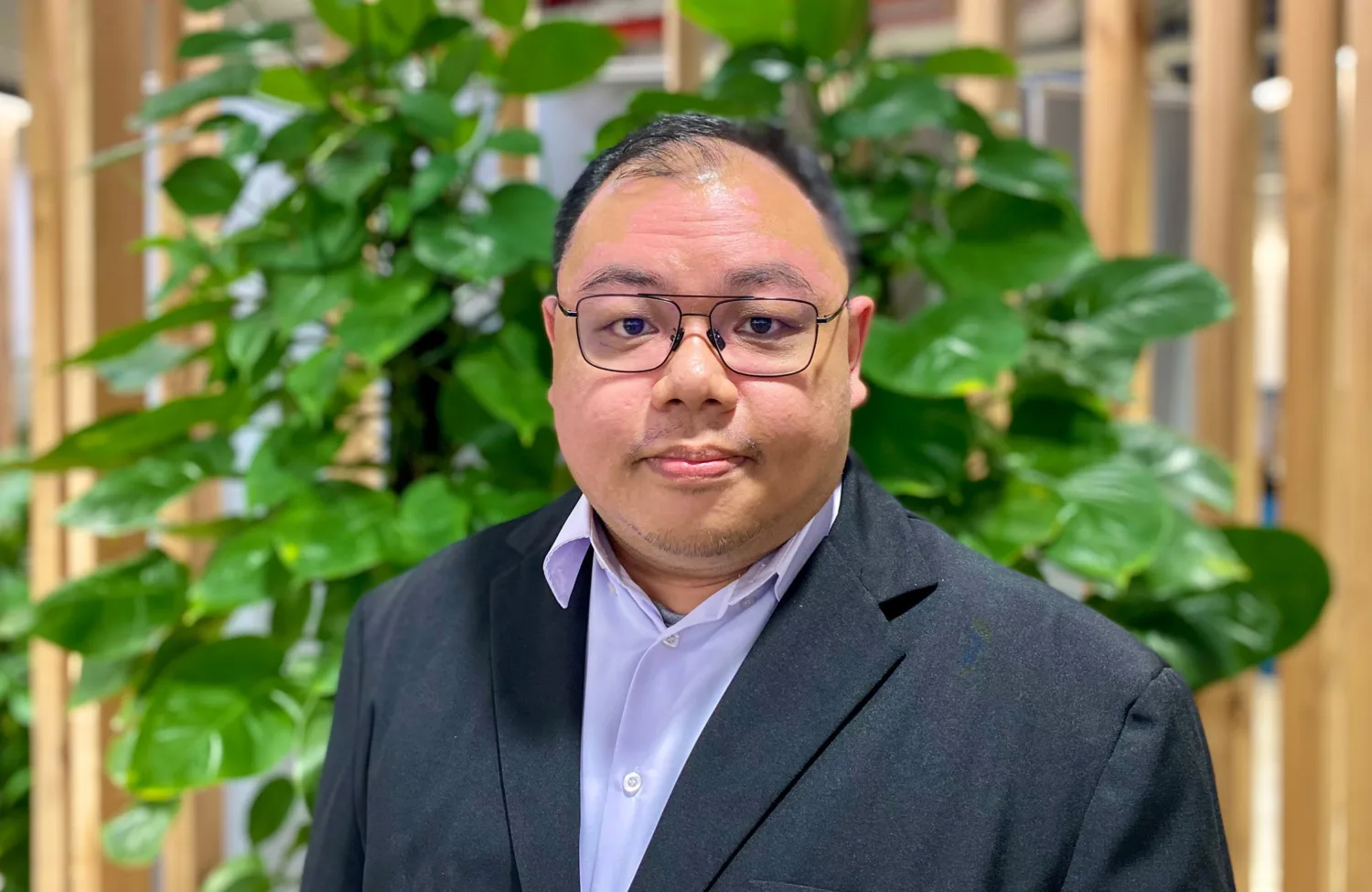 Man wearing a dark suit jacket and light dress shirt stands in front of a wooden slat wall with green leafy plants. Red pipes and industrial elements are visible in the indoor background.
