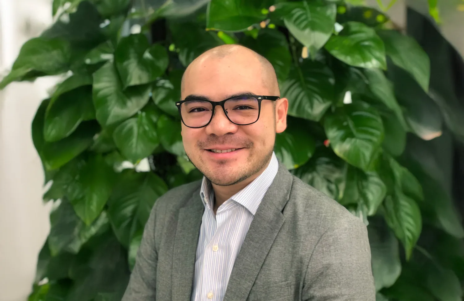 A person wearing glasses, a grey blazer, and a striped shirt is seated in front of a backdrop of dense green plants.