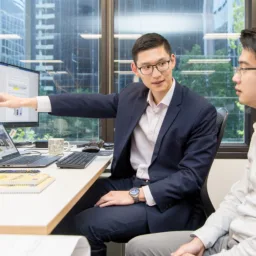 Alex sat in front of two computer screens pointing and discussing with a colleague