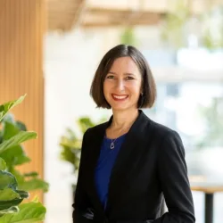 Annelies in a brightly lit open plan office, in a dark suit and blue top smiling to camera.