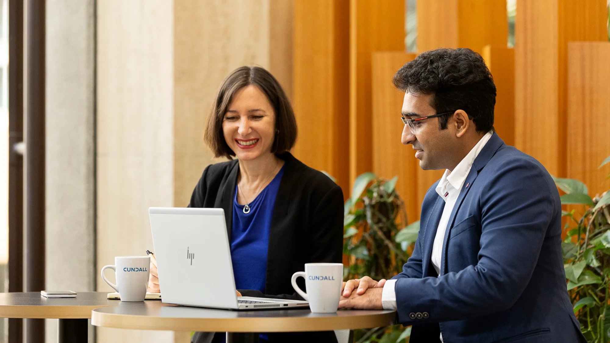 Annelies sat at a table with a colleague smiling and looking at a laptop screen.