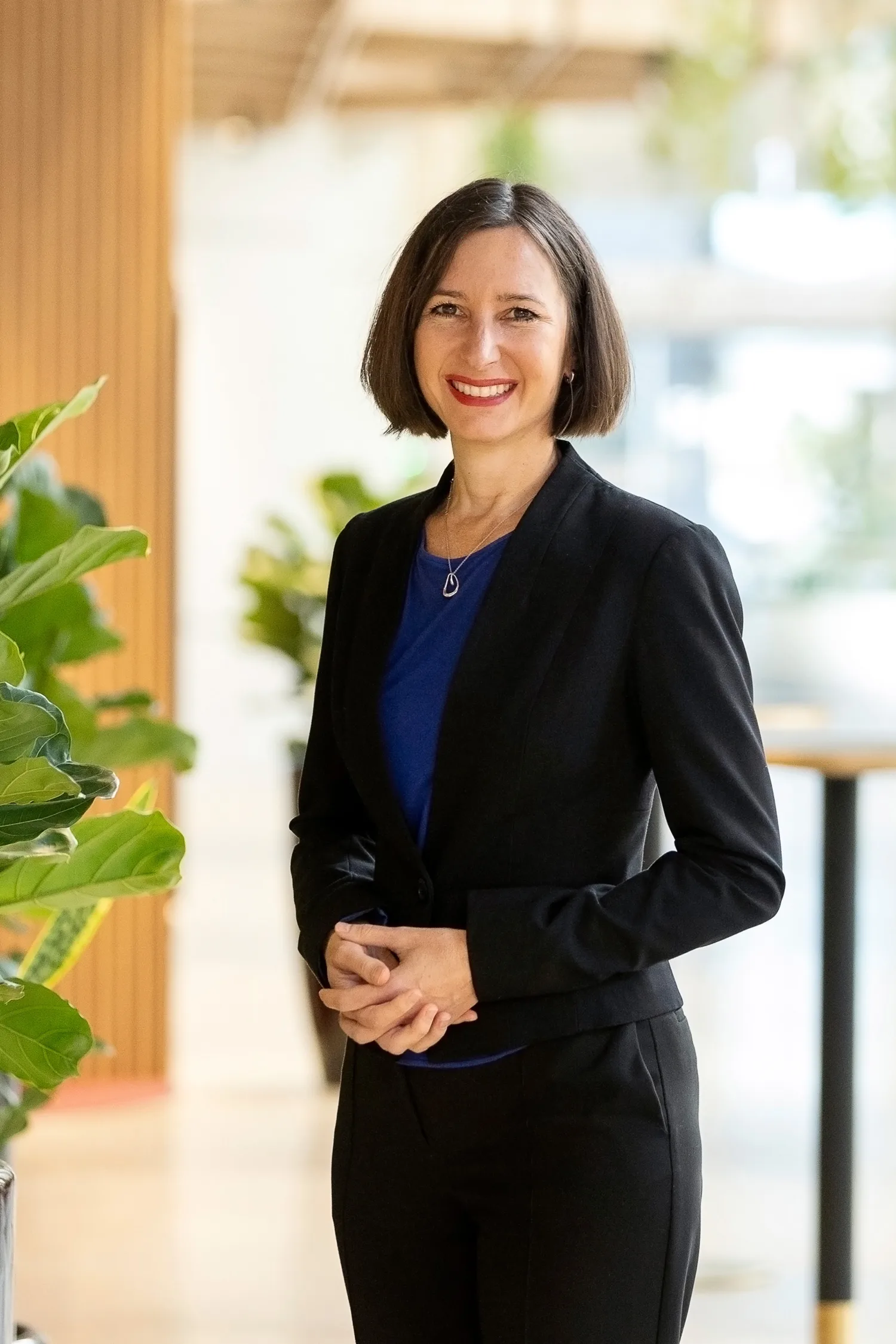 Annelies in a brightly lit open plan office, in a dark suit and blue top smiling to camera.