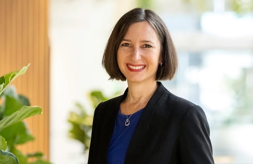 Annelies in a brightly lit open plan office, in a dark suit and blue top smiling to camera.