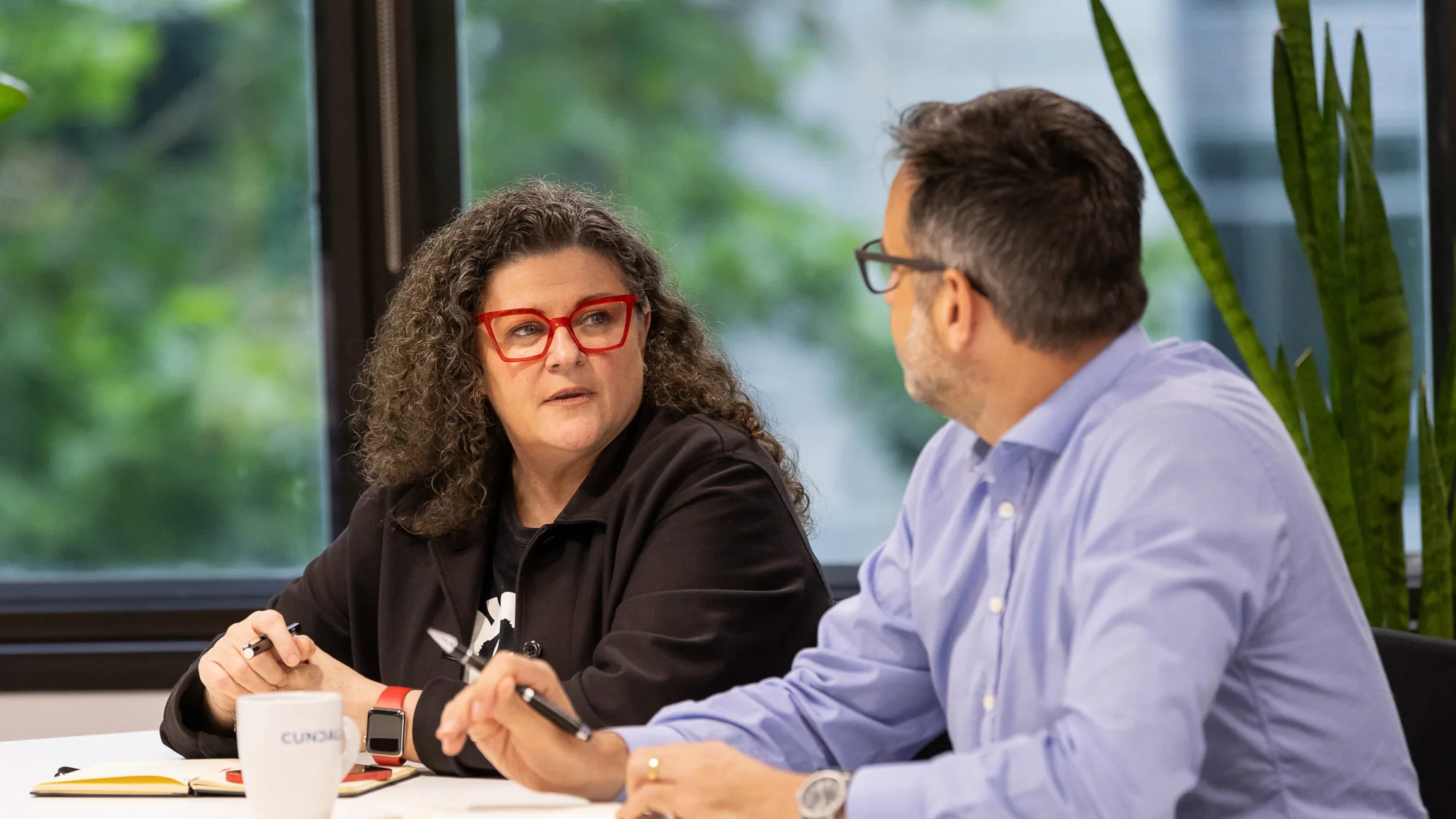 Bettina sat at an office table in conversation with a colleague with pens and notebooks