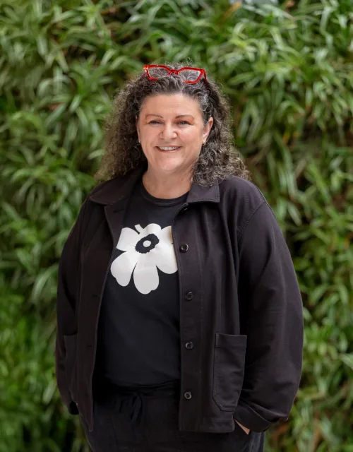 Bettina in dark clothing in front of a green hedge with red glasses on her head smiling to camera