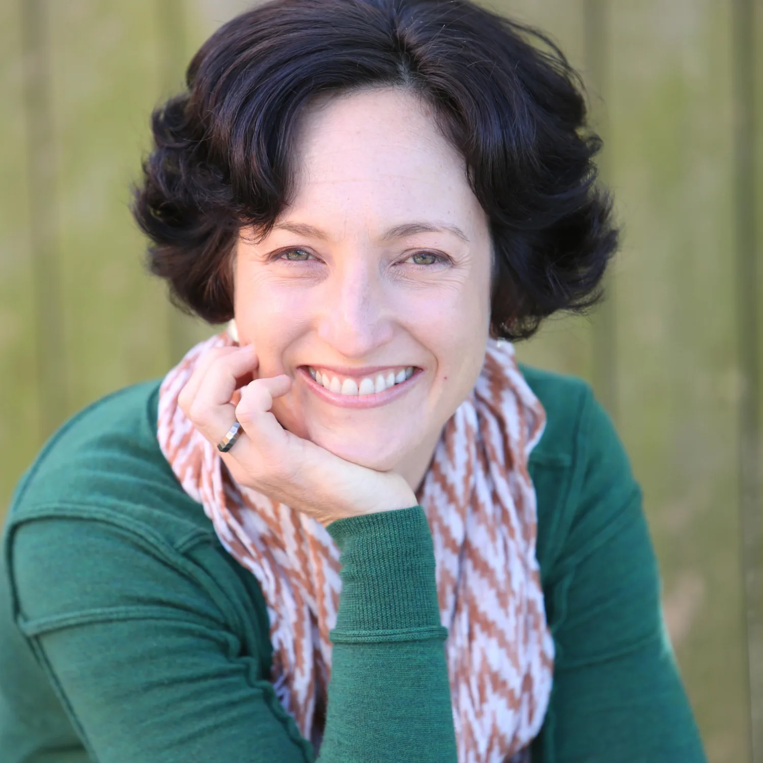 Jay Dee Dearness wearing a green long sleeved top and scarf, resting her head on her hand and smiling to camera