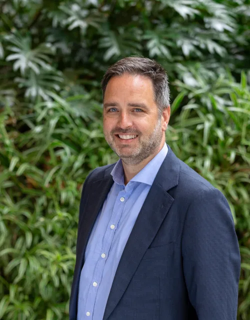 Julian, stood shoulder forward in a dark blue suit jacket and pale blue shite, smiling in front of a green hedge.