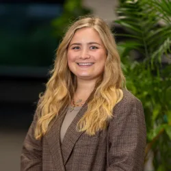 Katie Roberts wearing a brown plaid blazer over a beige top, standing indoors with hands clasped. Background includes green plants and blurred interior elements.