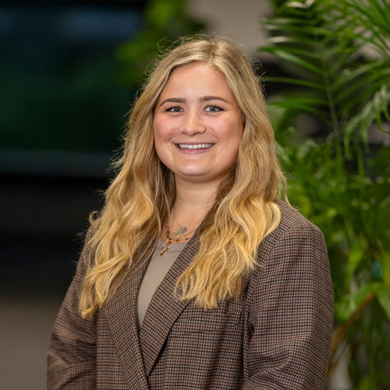 Katie Roberts wearing a brown plaid blazer over a beige top, standing indoors with hands clasped. Background includes green plants and blurred interior elements.