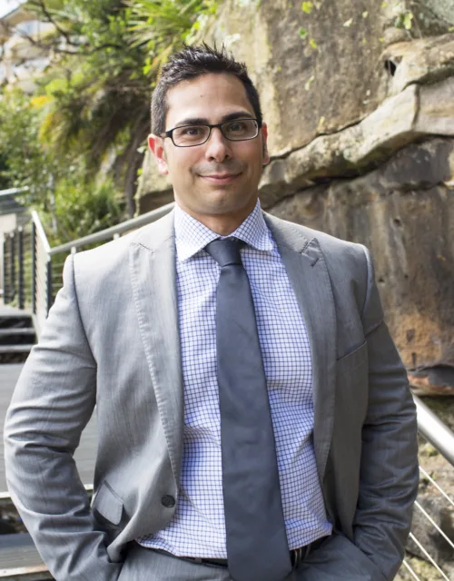 Mohammad Ali in a grey suit and blue shirt and tie with stairs and wall in the background
