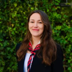 A person with long brown hair is standing outdoors in front of a dense green leafy background. They are wearing a black jacket over a white top and a red patterned scarf tied around the neck.
