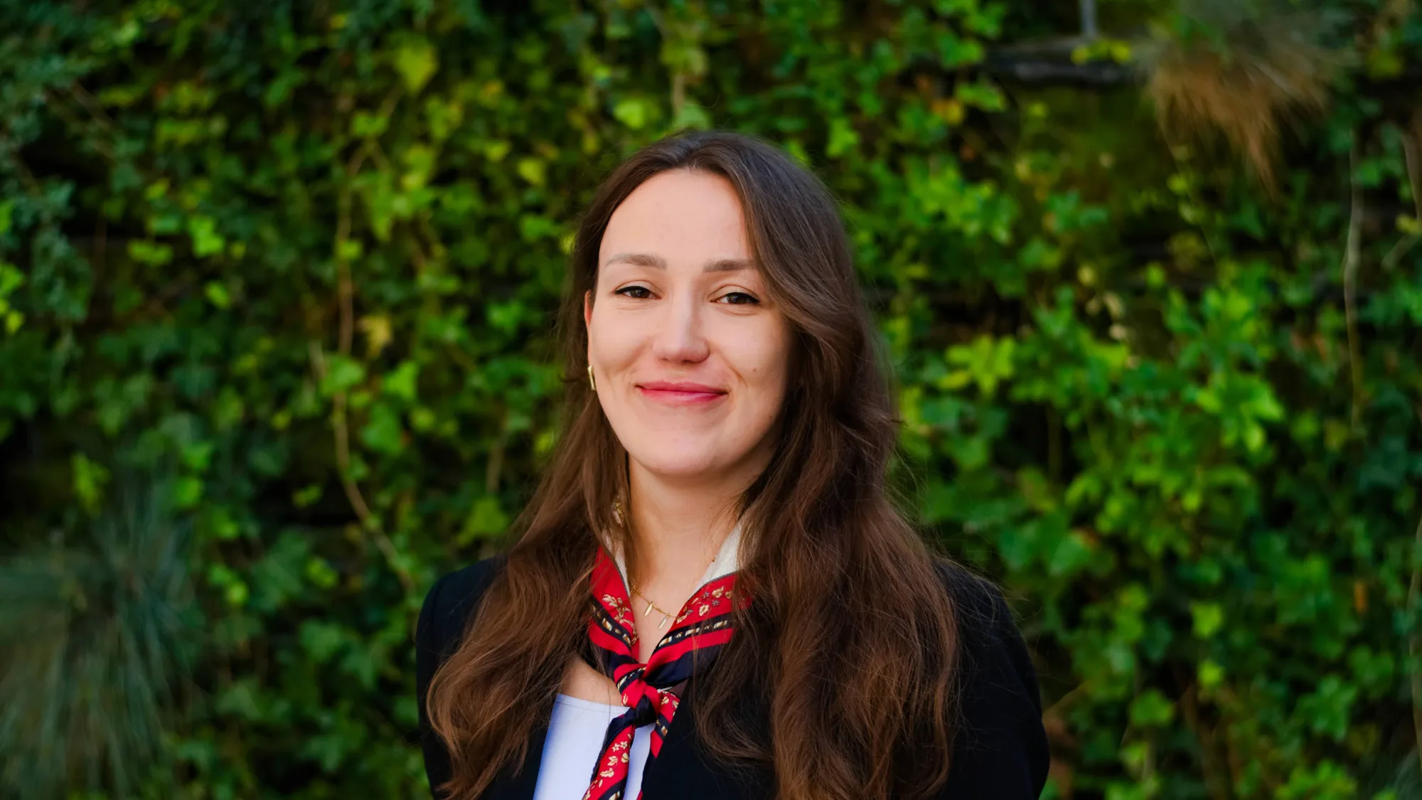 A person with long brown hair is standing outdoors in front of a dense green leafy background. They are wearing a black jacket over a white top and a red patterned scarf tied around the neck.