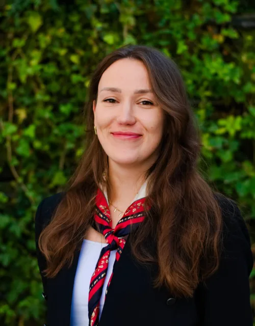 A person with long brown hair is standing outdoors in front of a dense green leafy background. They are wearing a black jacket over a white top and a red patterned scarf tied around the neck.