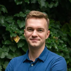 Andrzej Kaczmarek standing against a plain light-colored wall, wearing a light blue button-up shirt with a small collar and a black watch on the left wrist, arms crossed in front.