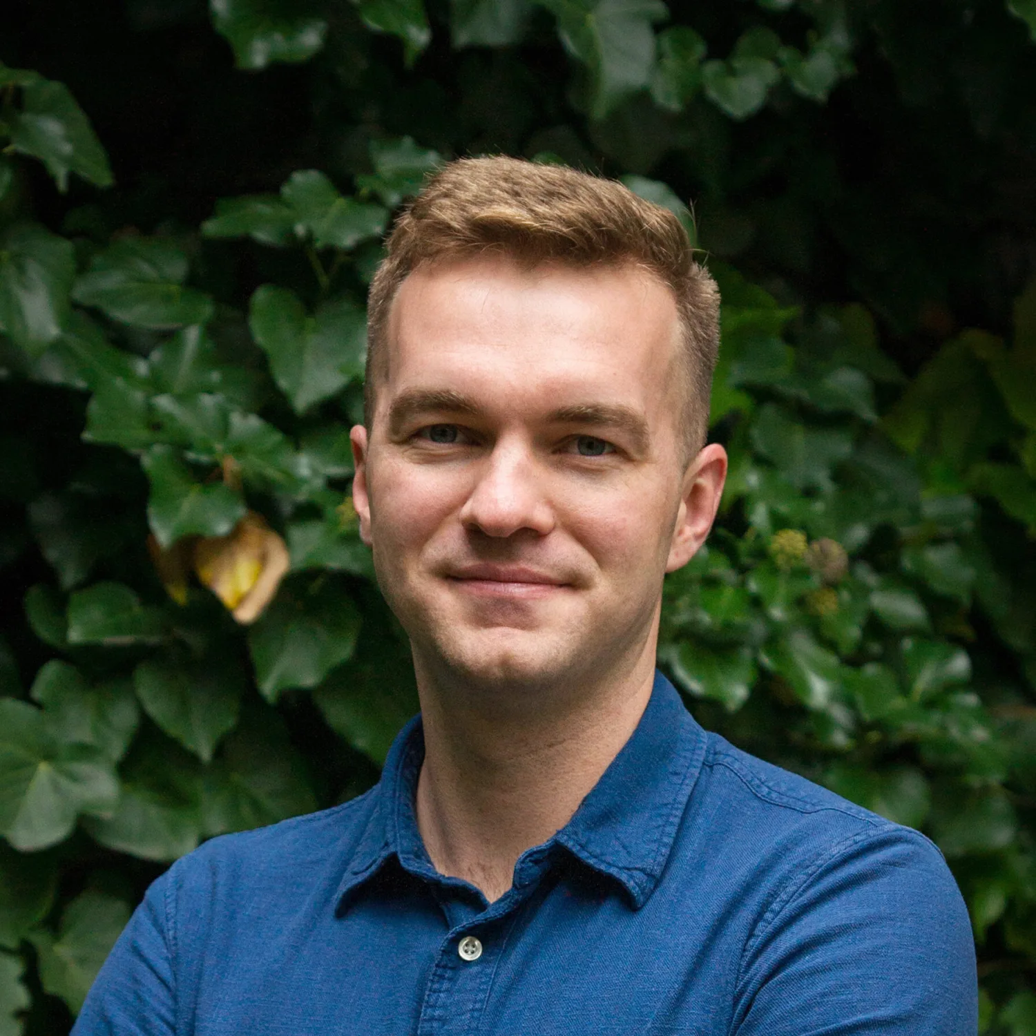 Andrzej Kaczmarek standing against a plain light-colored wall, wearing a light blue button-up shirt with a small collar and a black watch on the left wrist, arms crossed in front.