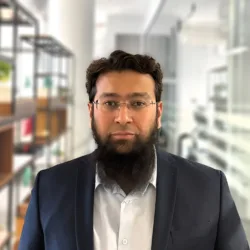 A person with glasses and a beard wears a dark suit jacket over a light shirt, standing in an office with shelves behind them containing books, decorative items, and potted plants.