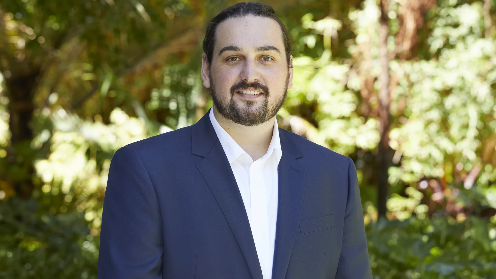 Head and sholders shot of Stefan wearing white shirt with blue blazer against a background of foliage