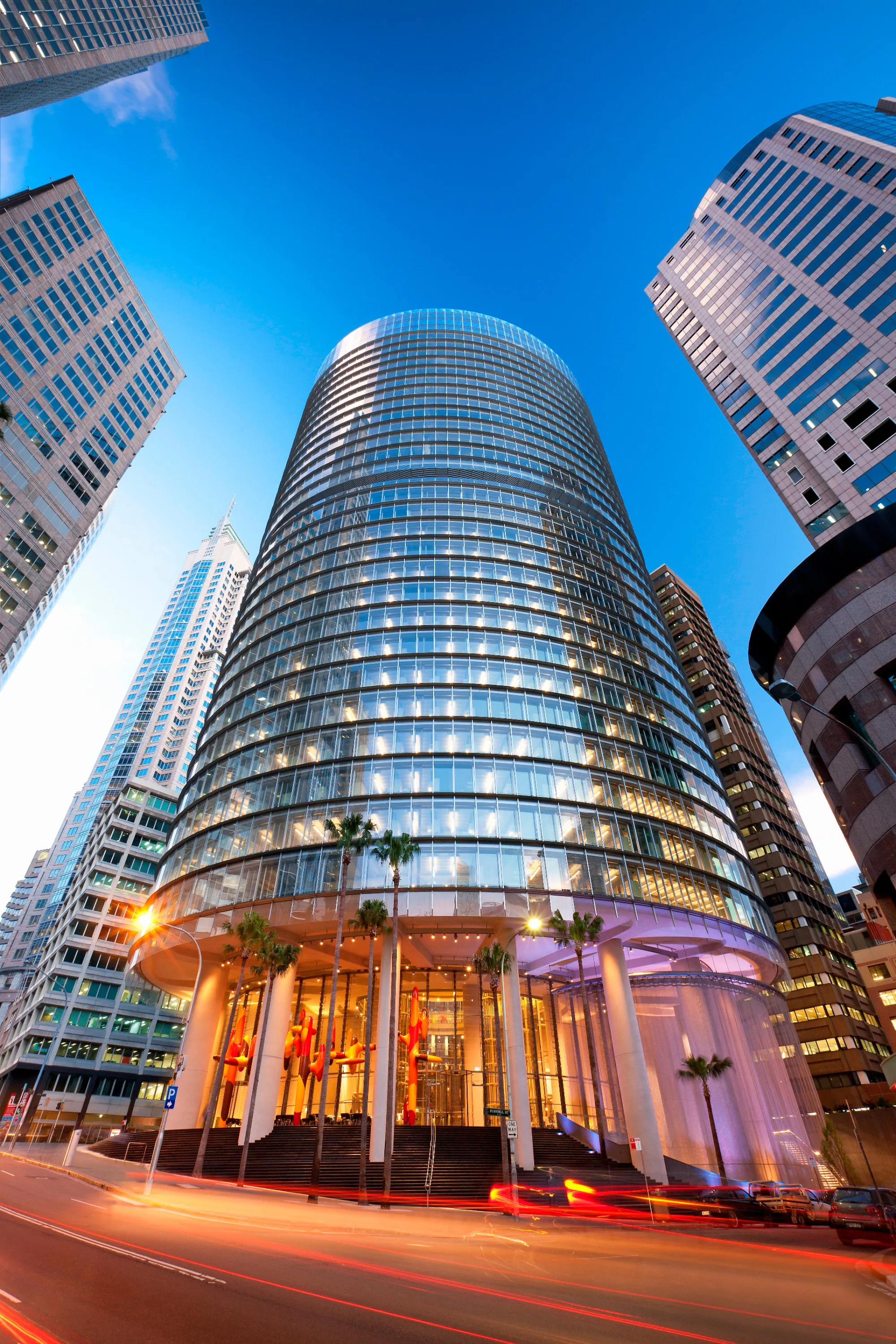 Tall glass skyscraper with curved façade and illuminated windows, surrounded by other high-rise buildings under a clear blue sky, with palm trees and light trails from vehicles at street level.