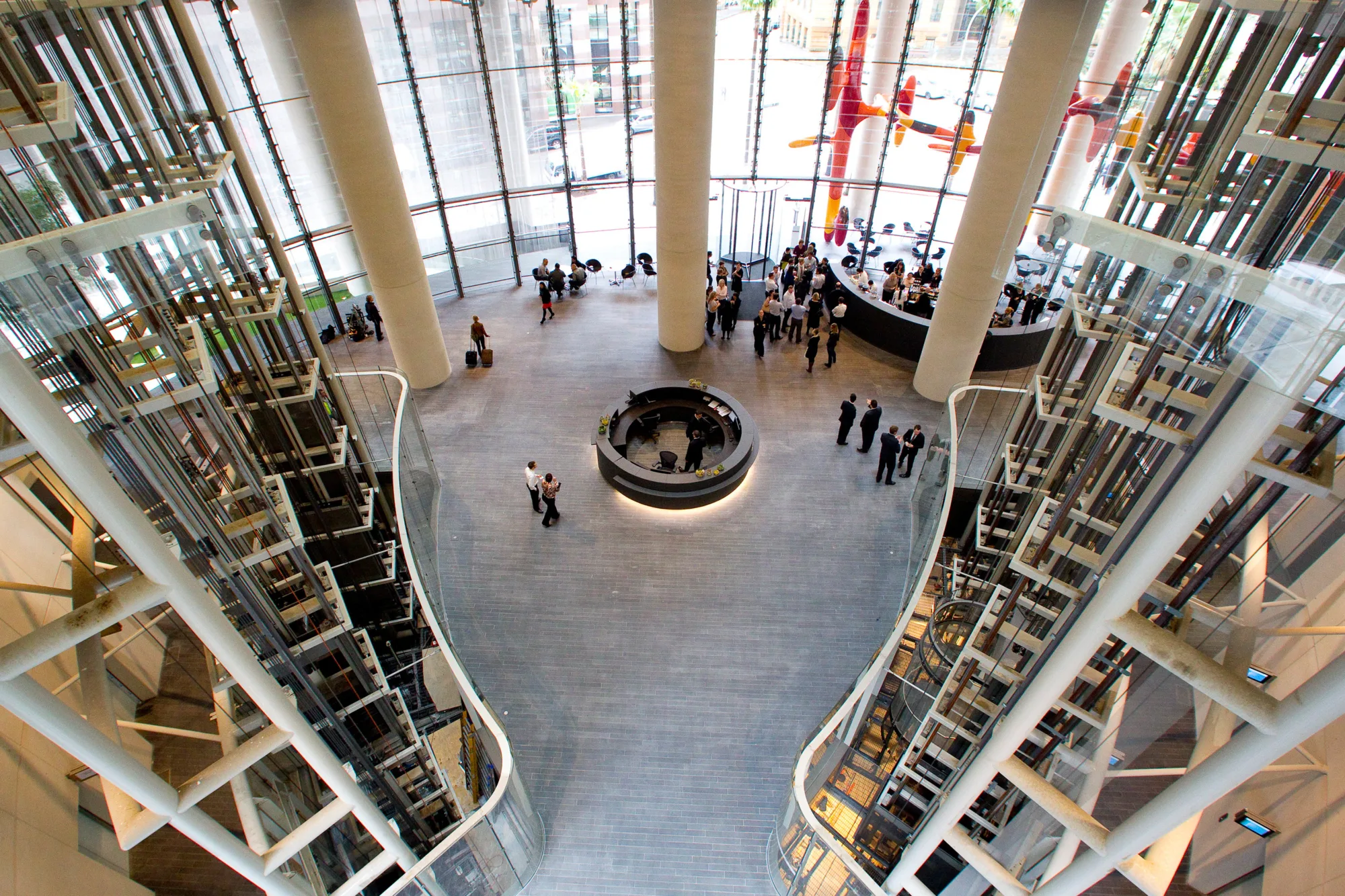 Interior view of a spacious lobby with tall glass walls, large vertical columns, and two glass elevator shafts on each side. A circular reception desk is centred on a grey-tiled floor, with groups of people standing near the desk and along the perimeter.