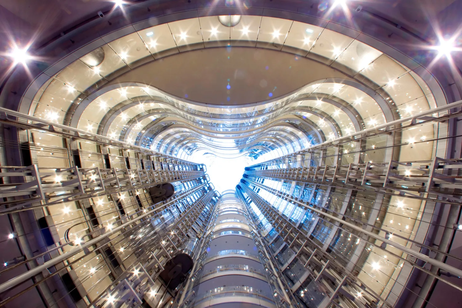 Interior view looking upward through a multi-storey atrium with curved glass walls, metal framework, and bright ceiling lights, converging towards a skylight at the top.