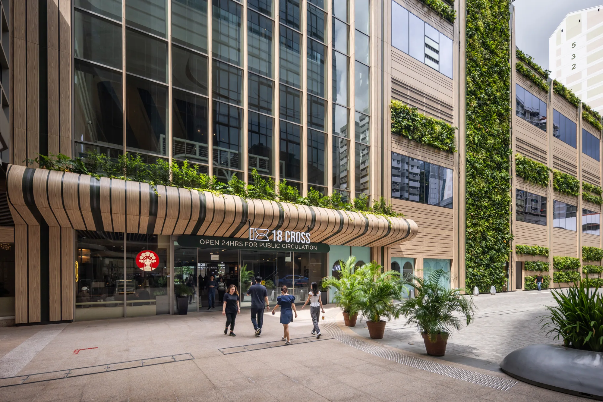 Exterior of a 18 Cross building with vertical gardens and large glass windows. Several people walk near the entrance, surrounded by potted plants.