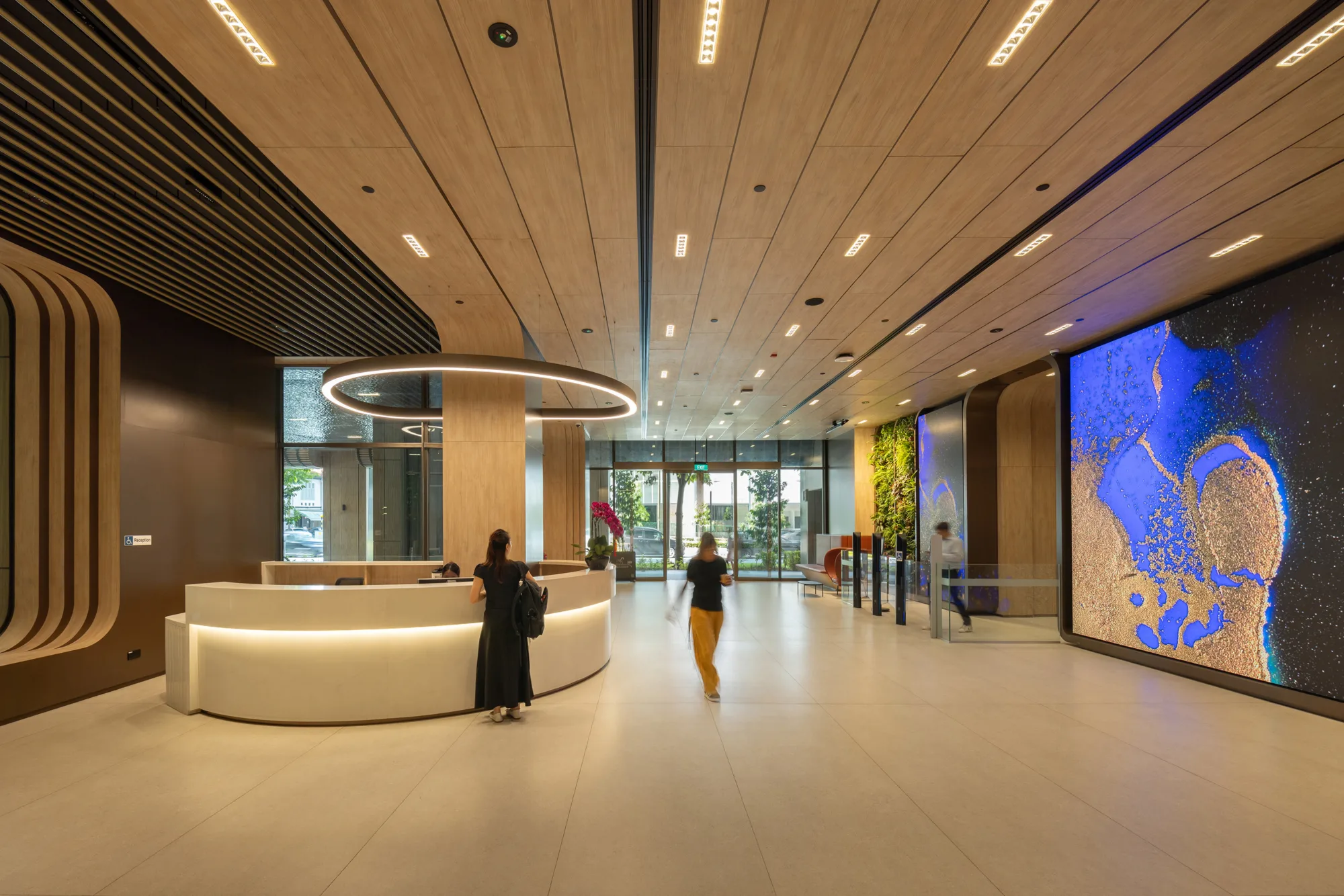 Interior of a 18 Cross building lobby with wood-paneled walls and ceiling. A circular light fixture hangs above the reception desk. Three people are present, a digital display screen fixture on the right. Glass doors and greenery are visible at the far end.