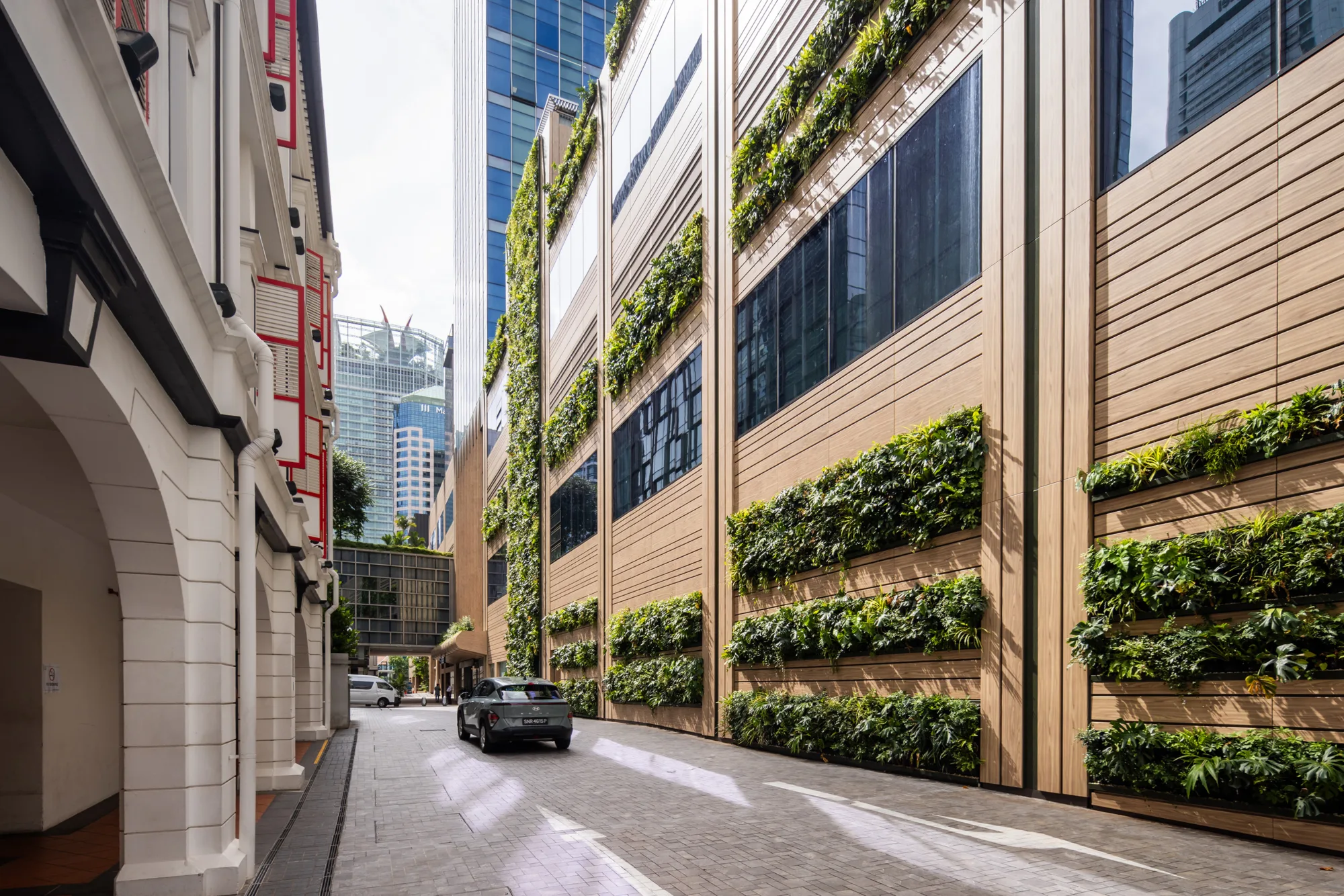 Narrow cobblestone street with buildings on both sides. The 18 Cross building on the right side features vertical gardens with plants in horizontal rows. A car is on the street, modern high-rise buildings appear in the background.