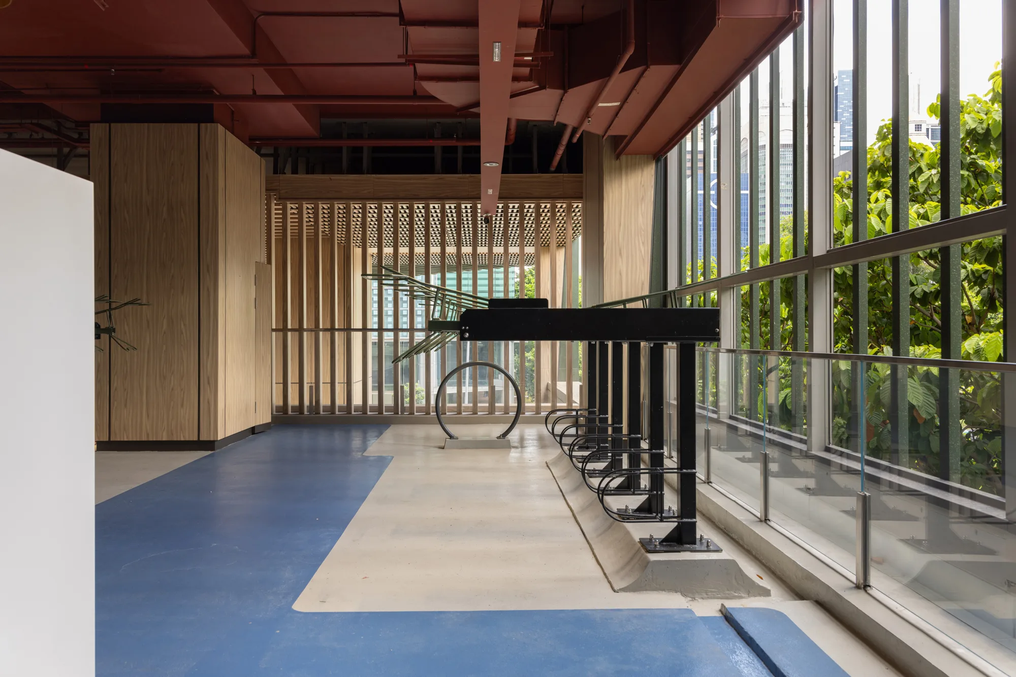 Indoor bike rack area with black metal racks on a blue and beige floor, glass windows on the right, red ceiling beams, and wooden panels on the left; greenery visible outside.