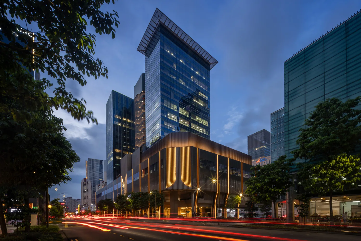 Cityscape at dusk featuring 18 Cross building. Light trails from traffic are visible on the busy street below, lined with trees and buildings nearby.