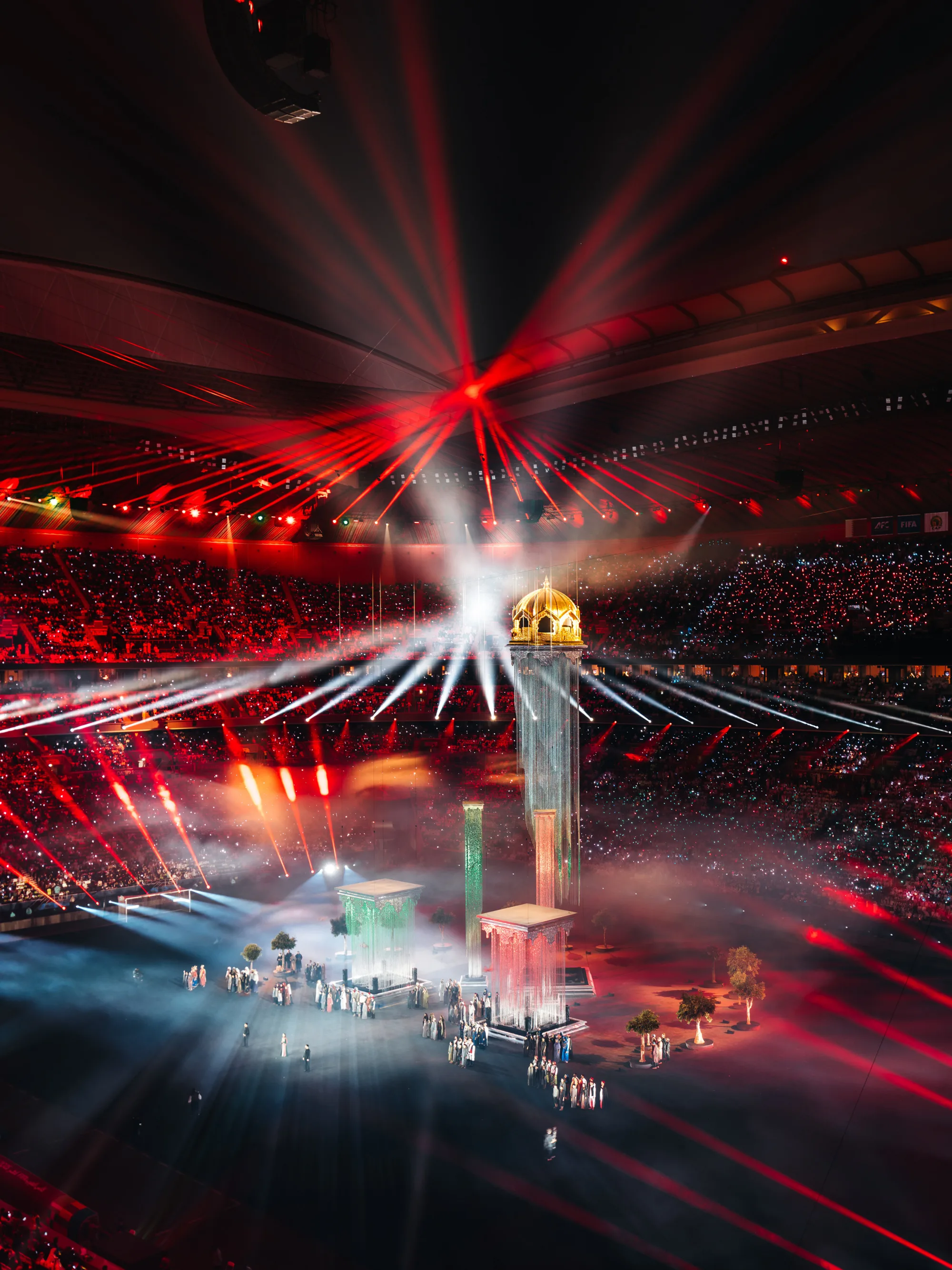 Interior of a large stadium with a central stage featuring tall column structures, one topped with a gold dome. Red and white laser lights radiate overhead, with groups of performers on the field and audience seating surrounding the scene.