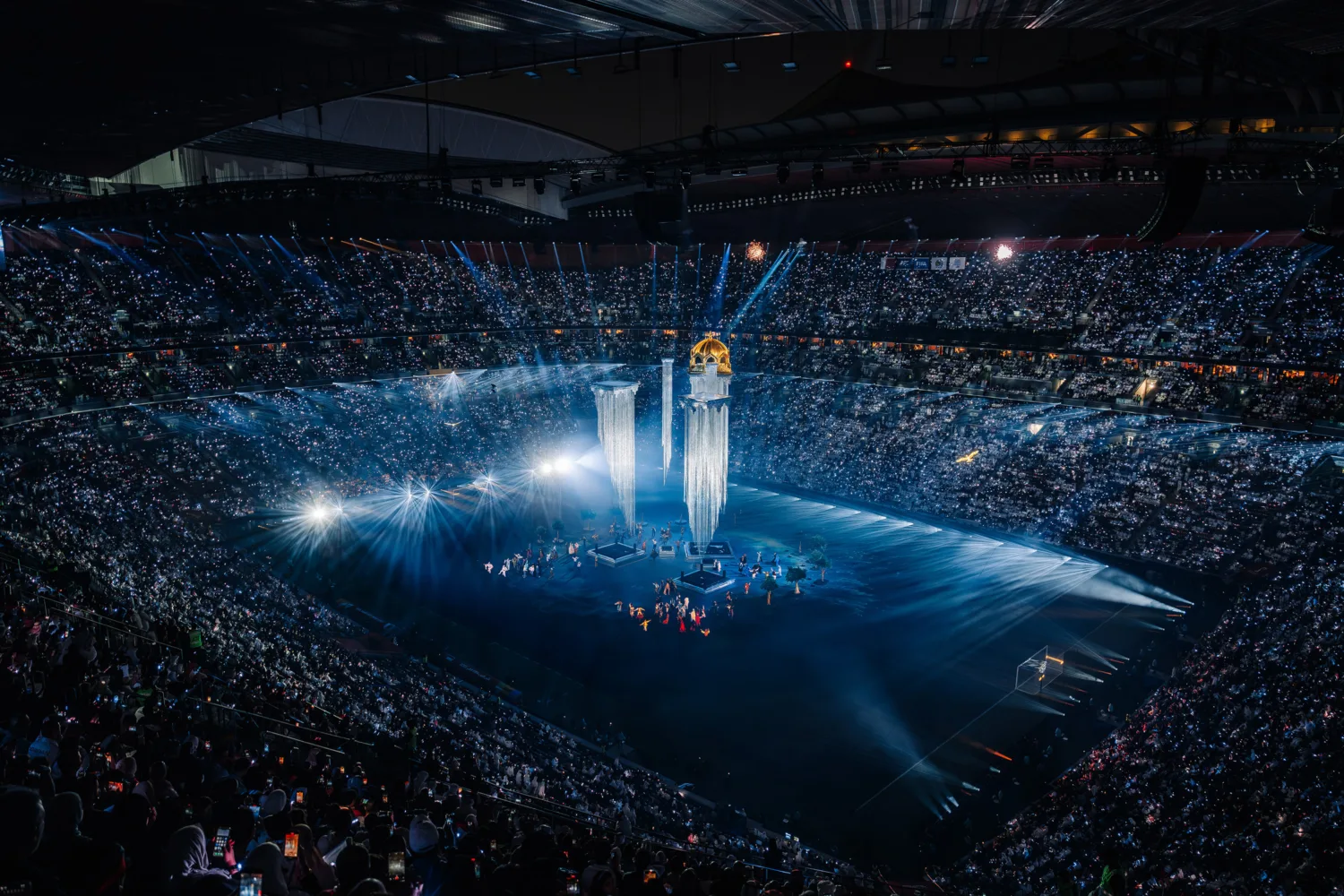 Wide view of a large stadium filled with spectators surrounding a central stage. Tall illuminated columns and lighting effects rise from the field, with spotlights and haze visible across the arena.
