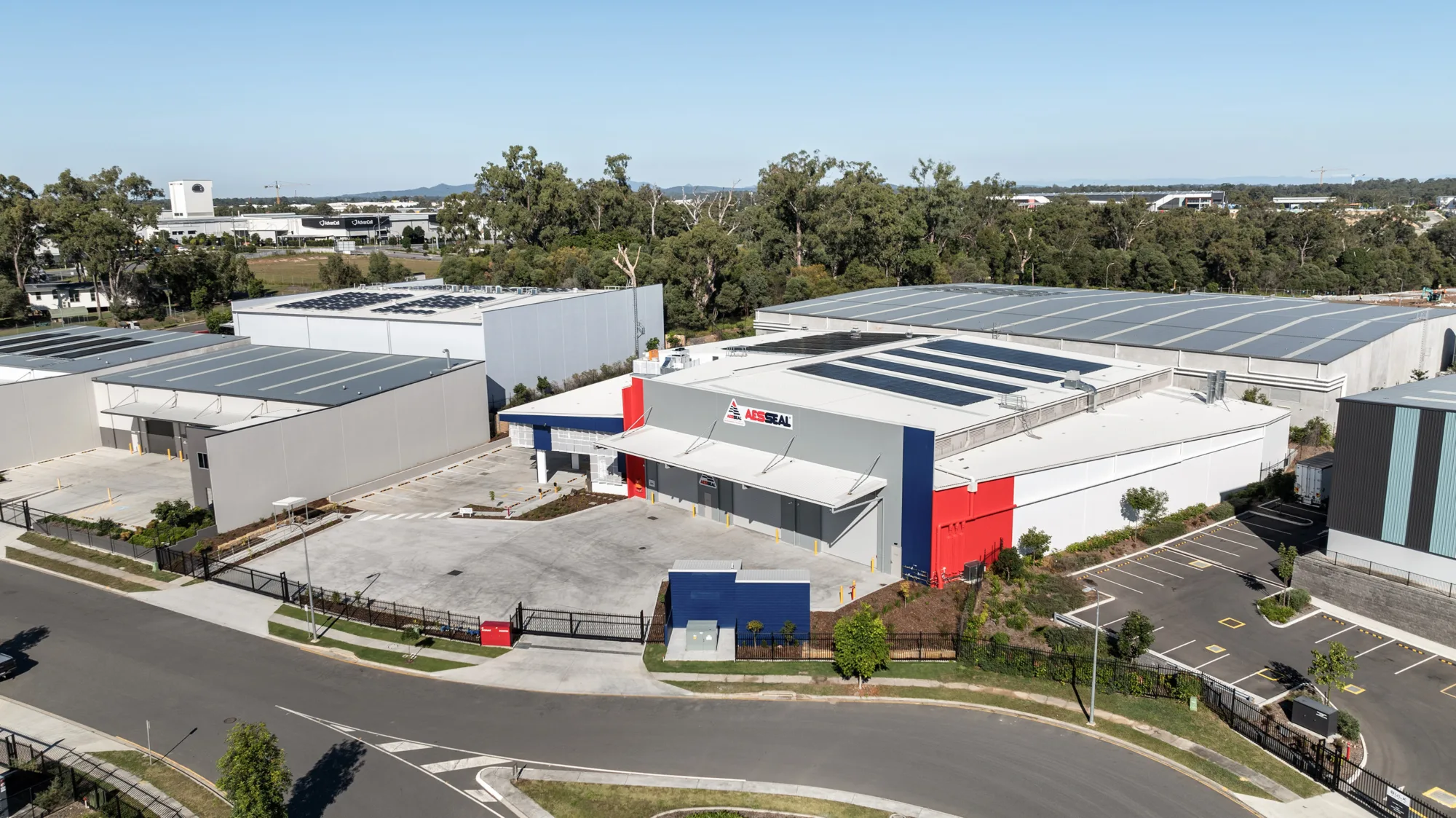 AESSEAL industrial facility aerial view showing grey warehouse buildings with solar panels, blue and red accents, paved driveway, gated entrance, and landscaped areas in a business park setting.