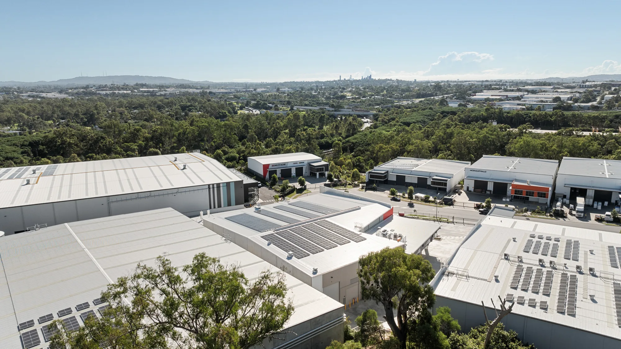 Aerial view of AESSEAL industrial complex with multiple grey warehouse roofs fitted with solar panels, surrounded by trees and roads, with distant city skyline in the background.