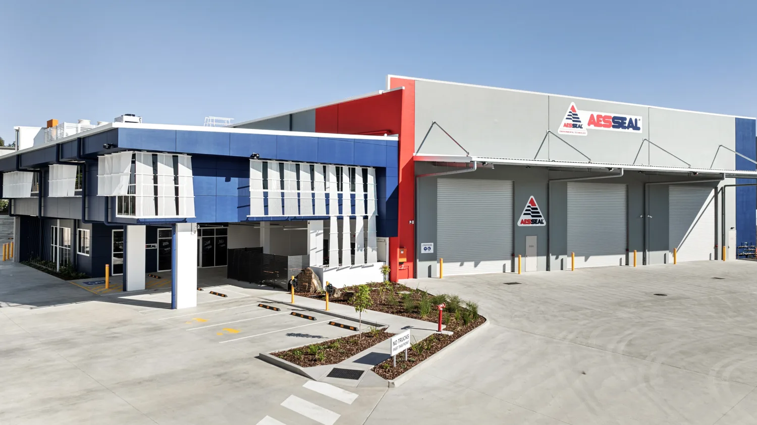 AESSEAL industrial facility exterior with blue and white office building and grey warehouse featuring three roller doors, AESSEAL logo signage, paved parking area, pedestrian crossings, and landscaped garden beds.