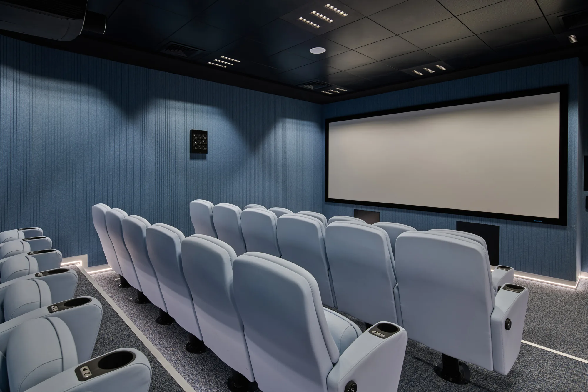 A cinema room with rows of cushioned reclining seats facing a large blank projection screen, surrounded by blue acoustic wall panels, recessed ceiling lights, and integrated speakers.